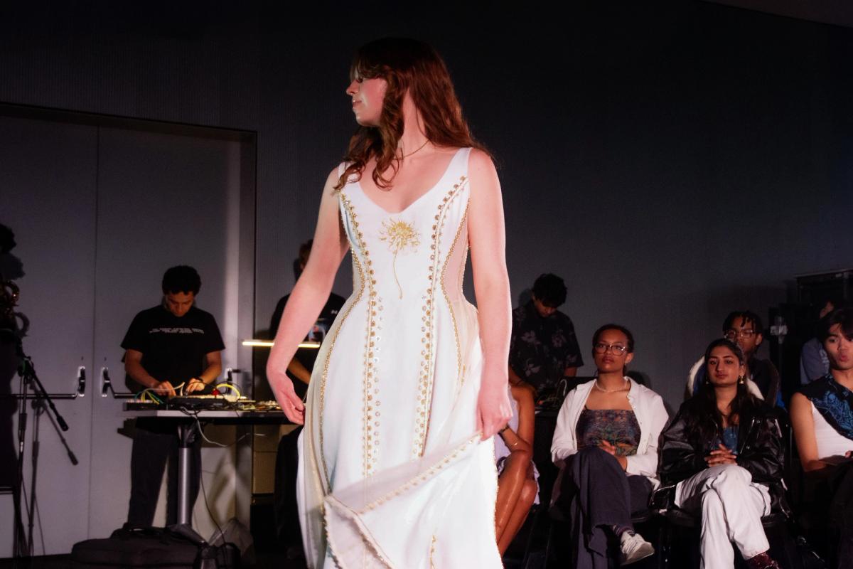 Model walks in a white dress featuring gold embroidery and a sunburst motif on the bodice. The structured gown has a sweeping hemline and is framed by seated spectators.